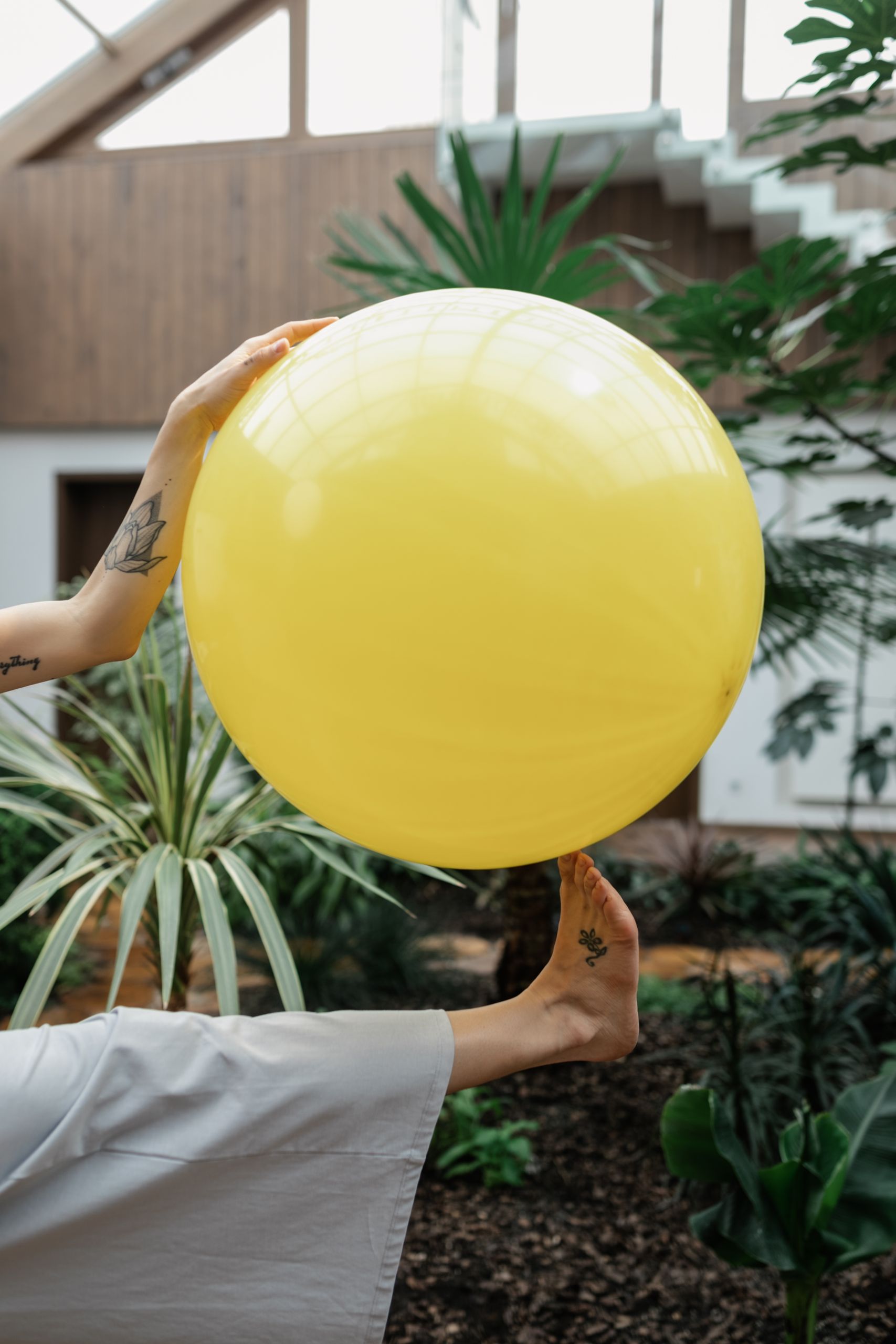 A yellow exercise ball held by a woman using her hands and feet in an indoor garden setting.